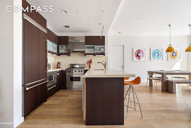 kitchen featuring built in appliances, wall chimney exhaust hood, a sink, a kitchen bar, and light countertops