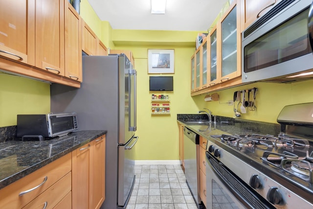 kitchen with appliances with stainless steel finishes, baseboards, a sink, glass insert cabinets, and dark stone counters