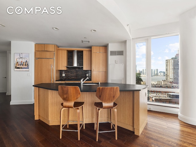 70 Little W St Unit 14B, New York City, NY 10004 - kitchen featuring wall chimney exhaust hood, dark countertops, modern cabinets, decorative backsplash, and dark wood-style floors