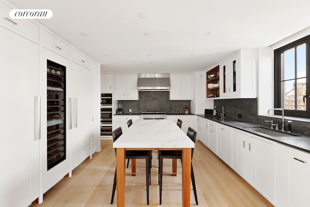 kitchen with a sink, wall chimney range hood, open shelves, backsplash, and light wood-style flooring