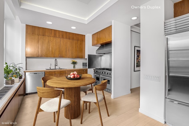 kitchen with high end appliances, a sink, wall chimney exhaust hood, recessed lighting, and brown cabinetry