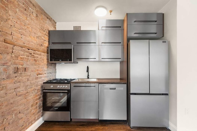 kitchen featuring stainless steel appliances, a sink, brick wall, baseboards, and gray cabinetry