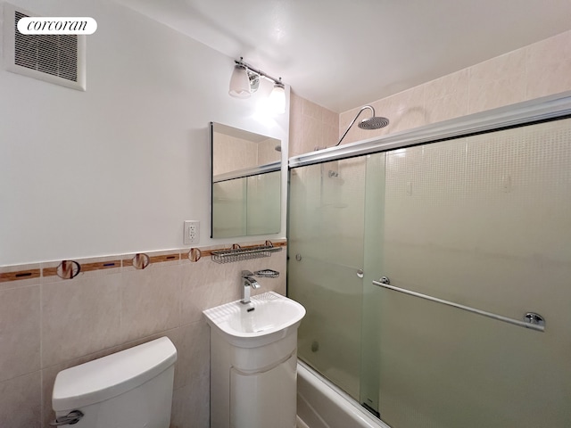 bathroom featuring toilet, tile walls, enclosed tub / shower combo, a sink, and wainscoting