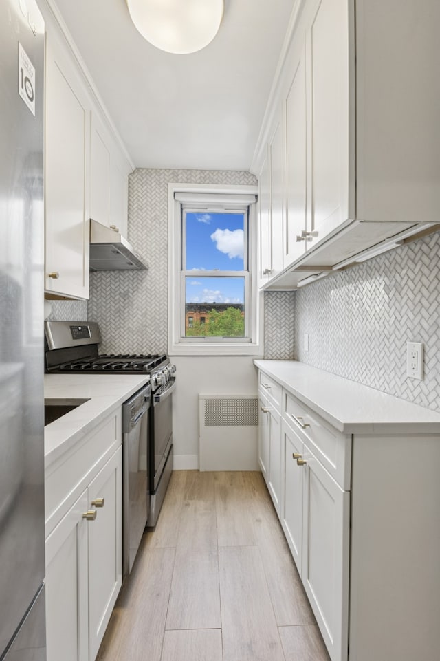 kitchen featuring appliances with stainless steel finishes, radiator heating unit, exhaust hood, light wood-style floors, and white cabinetry