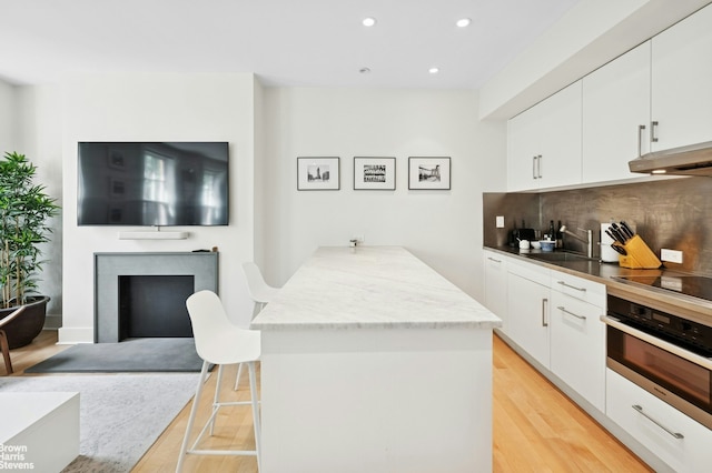 kitchen with stainless steel oven, black electric stovetop, light wood finished floors, backsplash, and white cabinetry