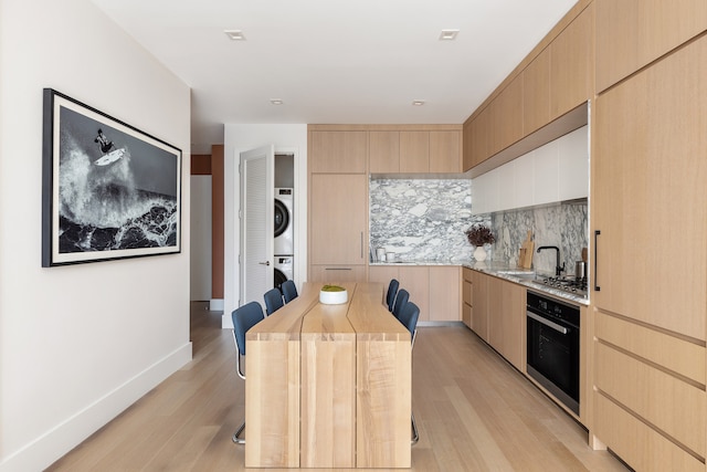 kitchen featuring light brown cabinets, oven, stacked washer / dryer, tasteful backsplash, and light wood-style floors