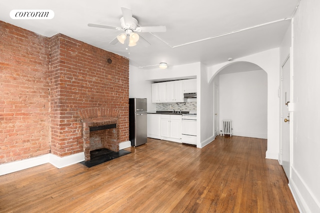 unfurnished living room with radiator heating unit, a ceiling fan, arched walkways, a brick fireplace, and wood-type flooring