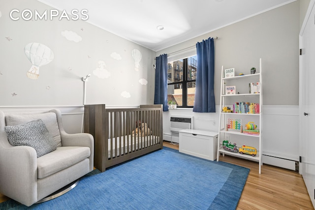bedroom with a nursery area, light wood-style flooring, ornamental molding, a wall mounted AC, and a baseboard radiator