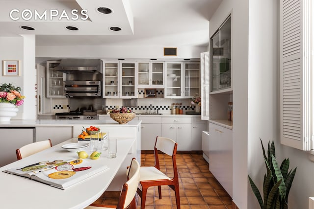 kitchen featuring decorative backsplash, light countertops, white cabinets, glass insert cabinets, and wall chimney range hood