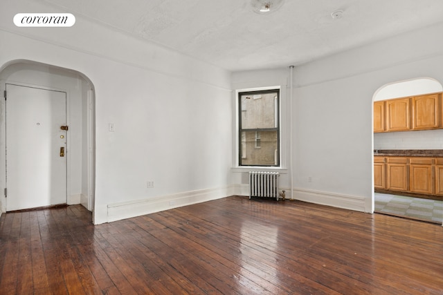 unfurnished living room featuring radiator heating unit, arched walkways, hardwood / wood-style flooring, and baseboards