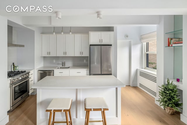 kitchen with appliances with stainless steel finishes, wall chimney range hood, a sink, light wood-type flooring, and a breakfast bar