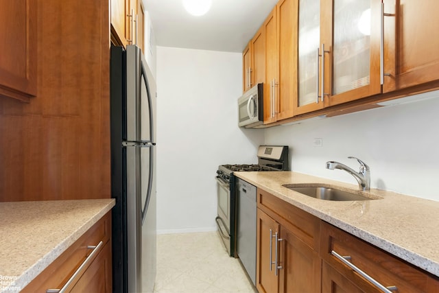 kitchen with a sink, appliances with stainless steel finishes, brown cabinetry, glass insert cabinets, and light stone countertops