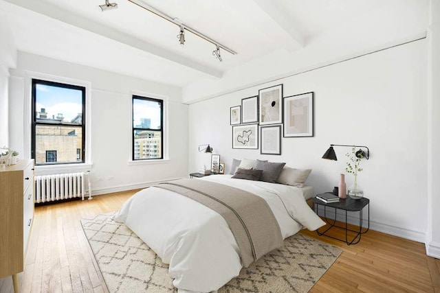 bedroom with radiator heating unit, rail lighting, light wood-style floors, beamed ceiling, and baseboards