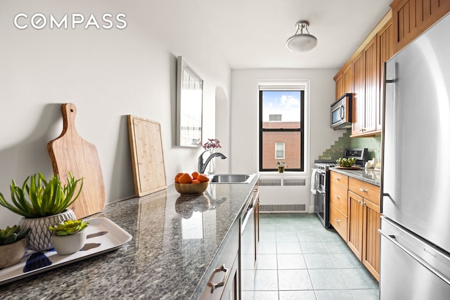 kitchen with appliances with stainless steel finishes, a sink, radiator, brown cabinetry, and dark stone countertops