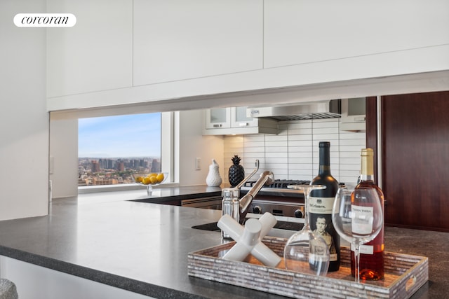kitchen view of backsplash, dark countertops, wall chimney range hood, and glass insert cabinets