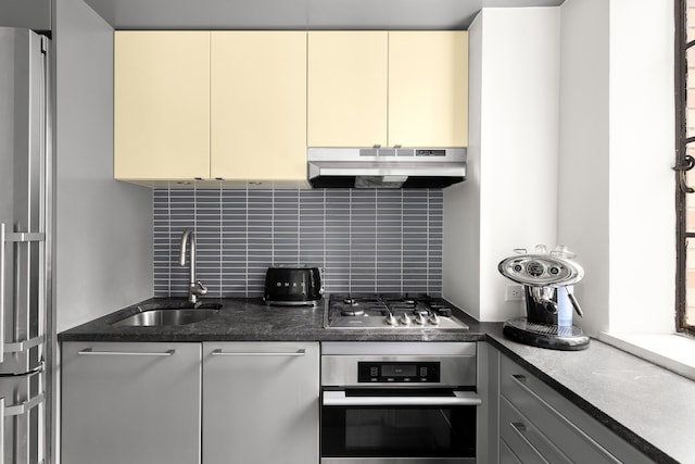 kitchen with stainless steel appliances, under cabinet range hood, a sink, cream cabinetry, and decorative backsplash