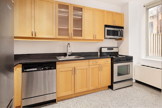 kitchen with appliances with stainless steel finishes, light brown cabinetry, a sink, dark countertops, and glass insert cabinets