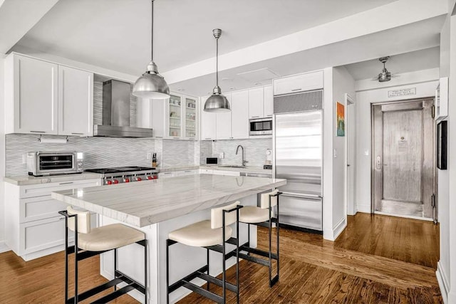 kitchen featuring wall chimney range hood, built in appliances, a sink, and decorative backsplash