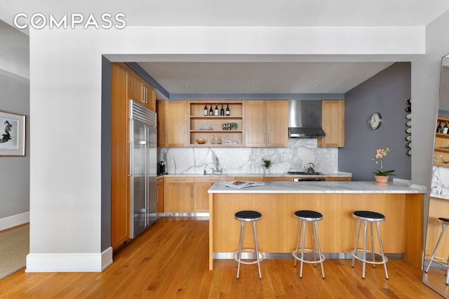 kitchen featuring built in refrigerator, wall chimney range hood, a peninsula, and light countertops
