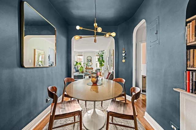 dining room featuring wood finished floors, arched walkways, a chandelier, electric panel, and baseboards