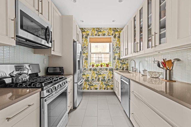 kitchen with appliances with stainless steel finishes, wallpapered walls, a sink, glass insert cabinets, and white cabinets