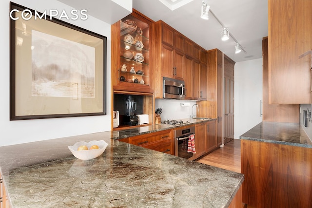 175 E Broadway Unit 8A, New York City, NY 10002 - kitchen with stainless steel appliances, a sink, brown cabinetry, dark stone countertops, and glass insert cabinets