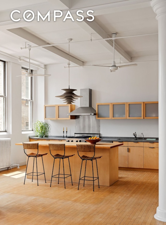 32 W 20th St Unit 9B, New York City, NY 10011 - kitchen with wall chimney range hood, beamed ceiling, radiator heating unit, light wood finished floors, and a kitchen breakfast bar