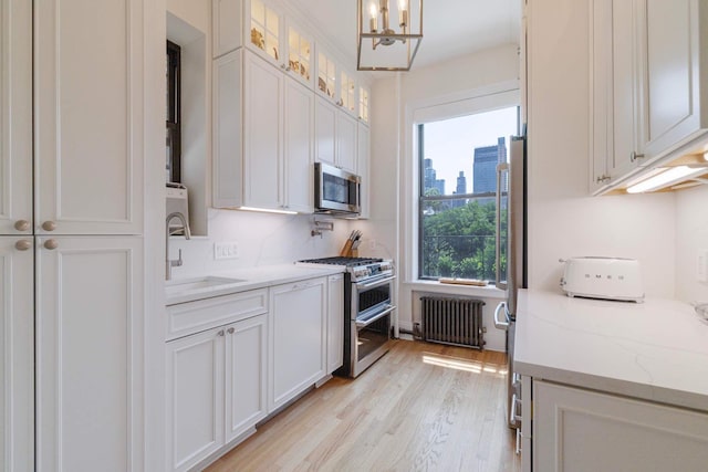 350 W 48th St Unit 4E, New York City, NY 10036 - kitchen with stainless steel appliances, radiator, a chandelier, white cabinetry, and light wood-style floors