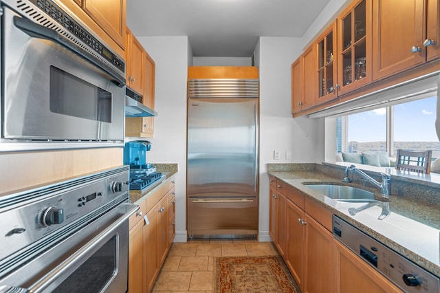 10 Little W St Unit 18-E, New York City, NY 10004 - kitchen with appliances with stainless steel finishes, under cabinet range hood, light stone counters, glass insert cabinets, and brown cabinetry