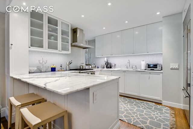 7 E 85th St Unit 5Ab, New York City, NY 10028 - kitchen with wall chimney exhaust hood, a breakfast bar, decorative backsplash, glass insert cabinets, and recessed lighting