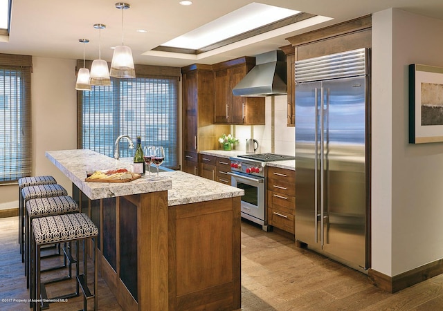 kitchen featuring baseboards, light wood finished floors, wall chimney exhaust hood, high quality appliances, and a center island with sink