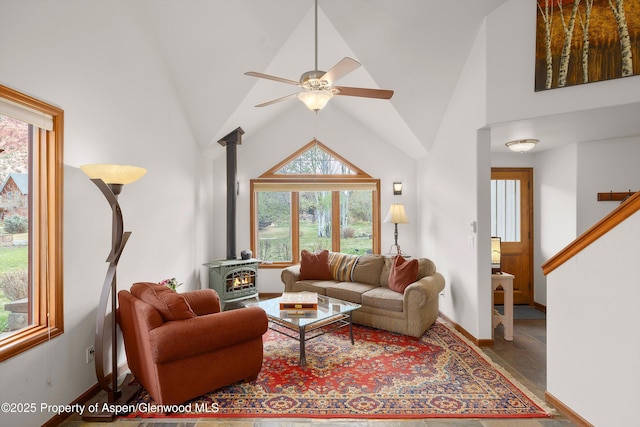 living room featuring a wood stove, baseboards, ceiling fan, stone finish floor, and high vaulted ceiling