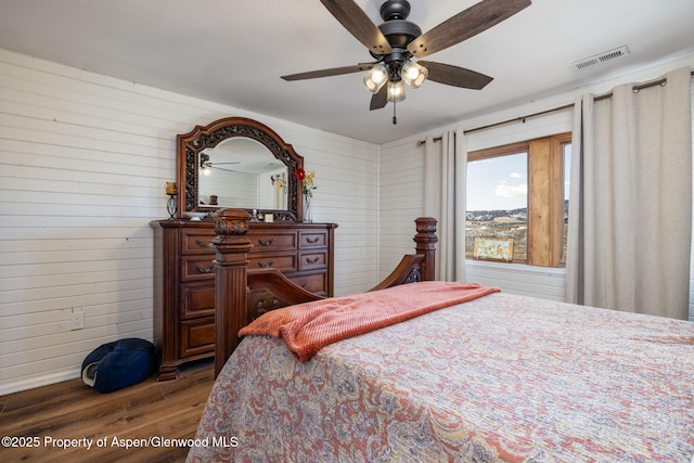 bedroom with a ceiling fan, wooden walls, visible vents, and dark wood-style flooring