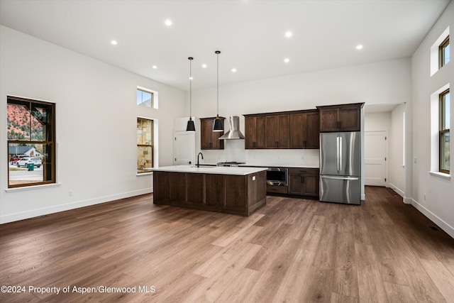 kitchen featuring pendant lighting, wall chimney range hood, stainless steel refrigerator, dark brown cabinetry, and an island with sink