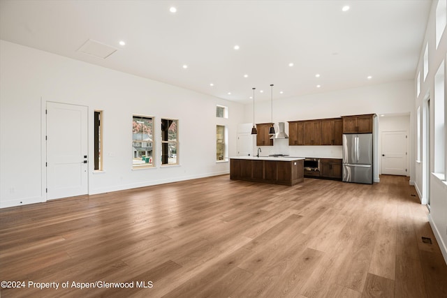 kitchen featuring an island with sink, stainless steel fridge, hanging light fixtures, wall chimney range hood, and light wood-type flooring