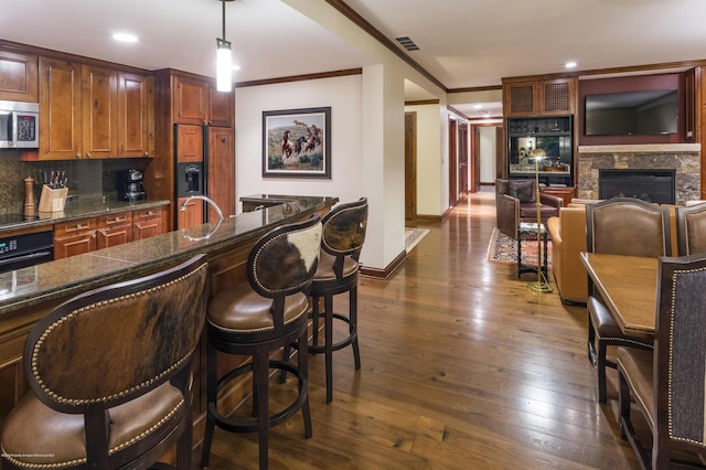 kitchen featuring dark wood-type flooring, hanging light fixtures, a stone fireplace, crown molding, and black appliances