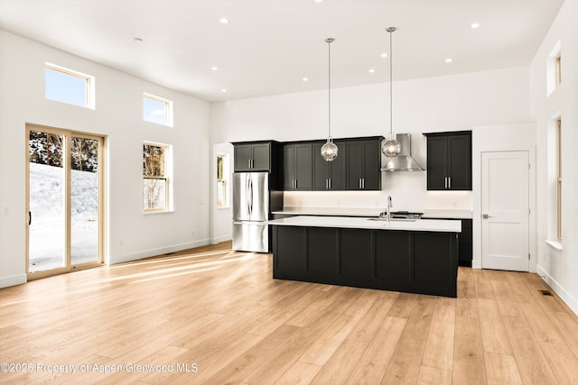 kitchen with a center island with sink, hanging light fixtures, stainless steel refrigerator, wall chimney range hood, and a high ceiling