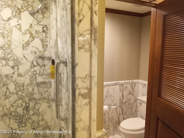 bathroom with ornamental molding, toilet, a marble finish shower, wainscoting, and tile walls