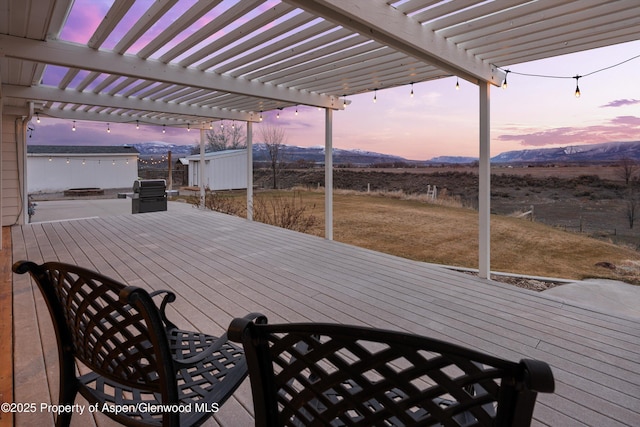 deck at dusk featuring a grill, a mountain view, a pergola, and an outdoor structure