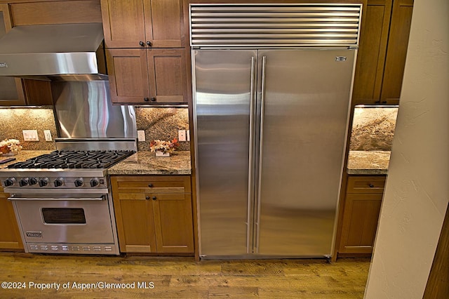 kitchen with wall chimney exhaust hood, high quality appliances, light wood-type flooring, and dark stone counters