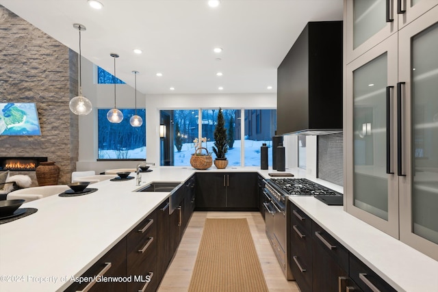 kitchen with a stone fireplace, wall chimney range hood, high end stainless steel range oven, sink, and hanging light fixtures