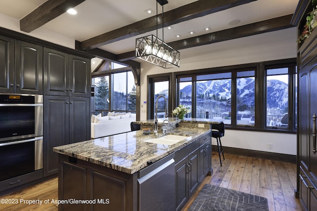 kitchen featuring light stone countertops, double oven, a center island with sink, a mountain view, and hanging light fixtures