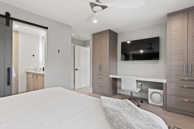 bedroom featuring connected bathroom, ceiling fan, a barn door, built in desk, and light wood-type flooring