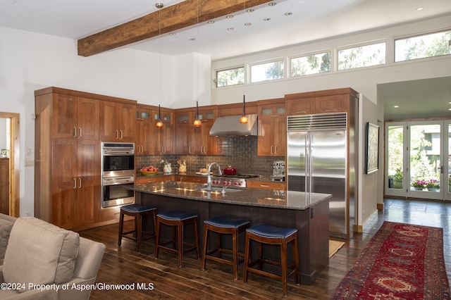 kitchen featuring dark stone counters, a kitchen island with sink, beam ceiling, a high ceiling, and range hood