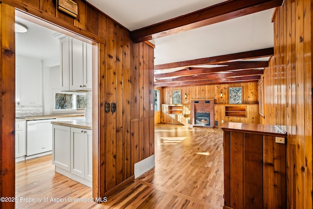 kitchen featuring a fireplace, white dishwasher, beam ceiling, white cabinets, and wood walls