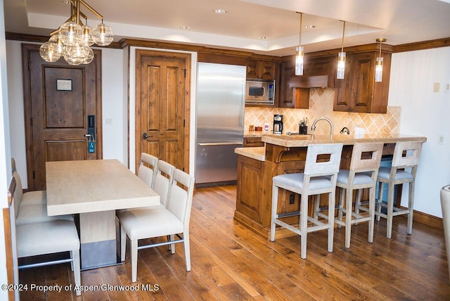 kitchen with a kitchen bar, light stone countertops, a tray ceiling, pendant lighting, and built in appliances