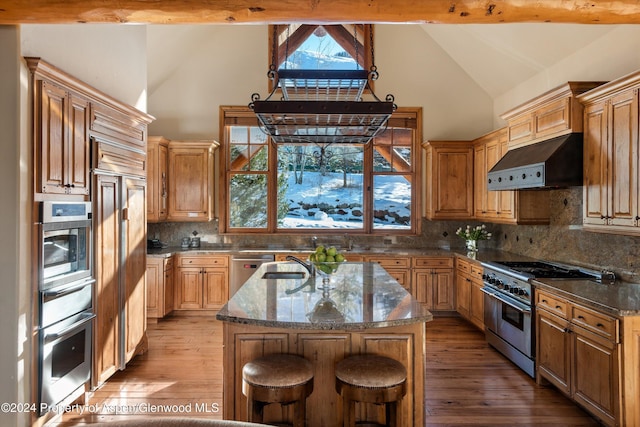 kitchen with tasteful backsplash, dark stone counters, stainless steel appliances, a kitchen island with sink, and beam ceiling