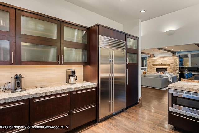 kitchen featuring a stone fireplace, dark brown cabinets, built in appliances, light hardwood / wood-style floors, and decorative backsplash