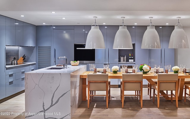 kitchen with a kitchen island with sink, sink, hanging light fixtures, light stone countertops, and light hardwood / wood-style floors