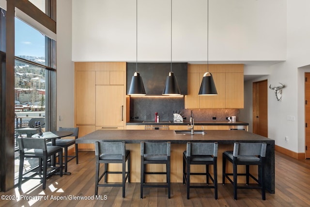 kitchen with light brown cabinetry, an island with sink, sink, decorative backsplash, and hanging light fixtures